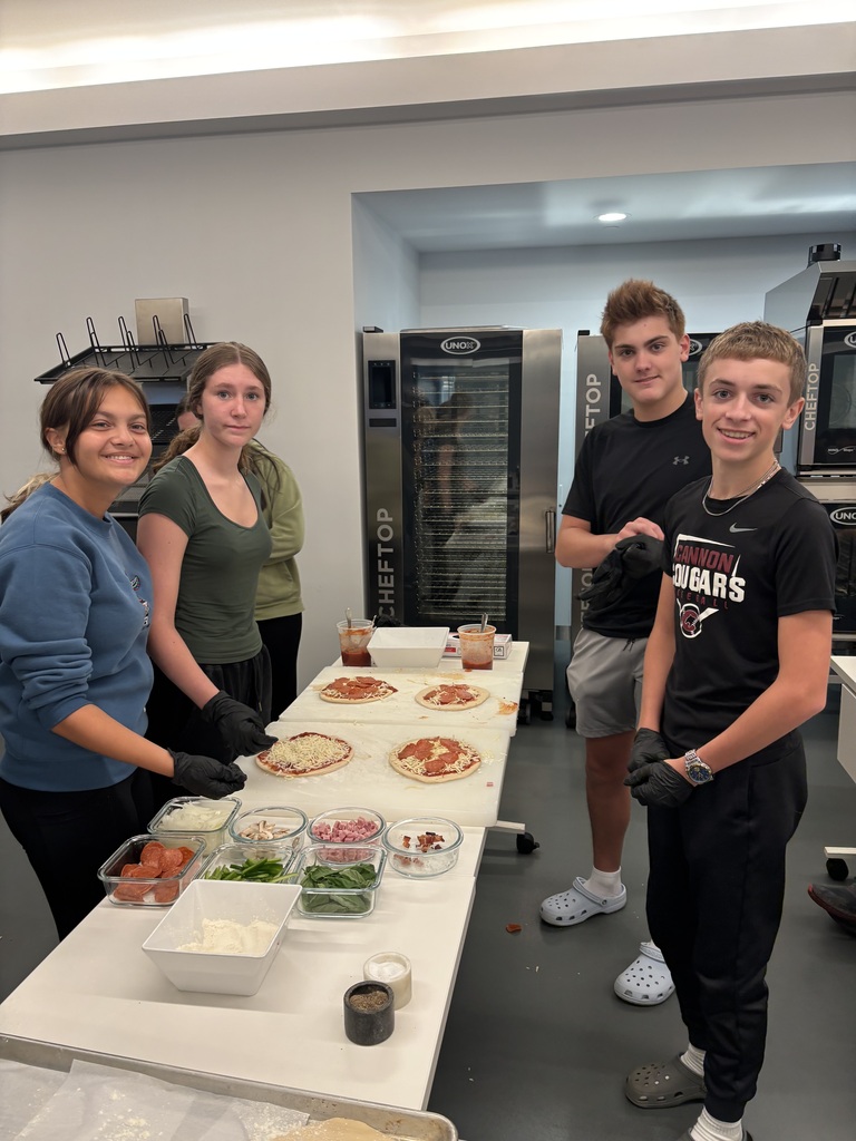 Four people in a commercial kitchen. They stand behind a long table with multiple pizzas and ingredients.
