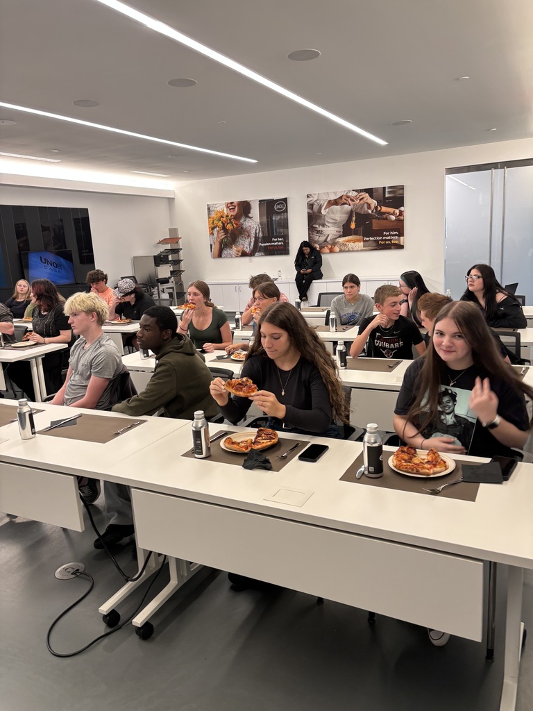 A group of people in a classroom are seated at tables, eating pizza. The room has white walls and pictures on them.