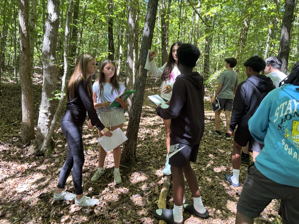 Several people are standing in a forest, with a woman holding paper and speaking to others.