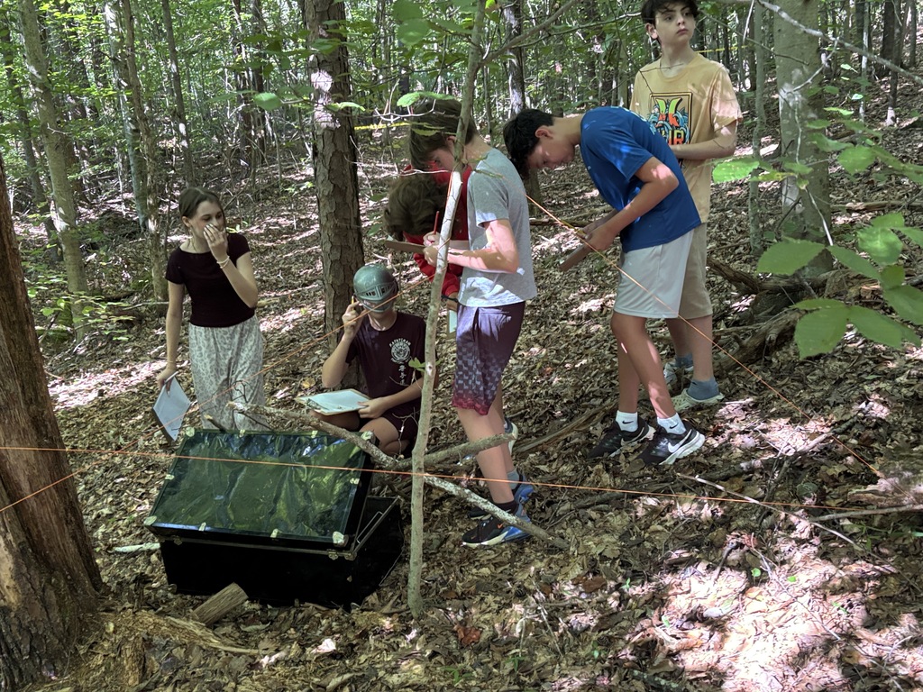 Five individuals stand in a forest, one seated, with two looking down and three standing. A black object with a green top is on the ground.