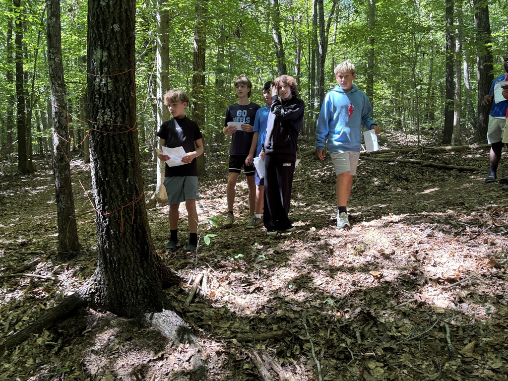 Several children in casual clothes gather in a dense forest, with some holding papers, one holding a map.