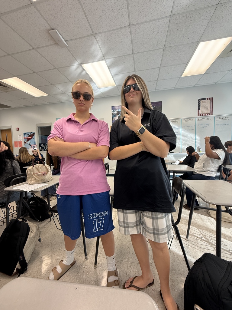 Two people stand in a classroom, one in a pink shirt and blue shorts, the other in black with sunglasses, both wearing sandals. Tables and chairs are behind them.