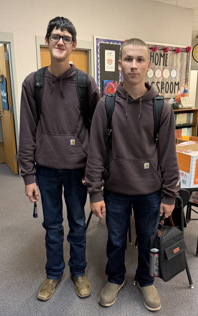 Two boys in matching brown hoodies and jeans stand in a classroom. One carries a backpack, the other a bag. A table and shelves are in the background.