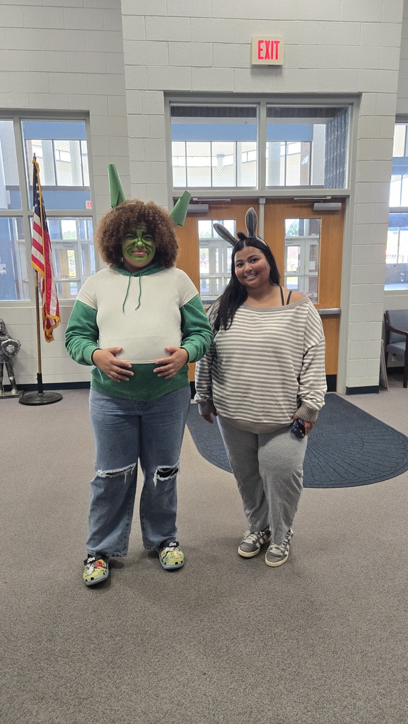 Two individuals stand inside a hallway. One is in a green costume. An American flag is behind them.