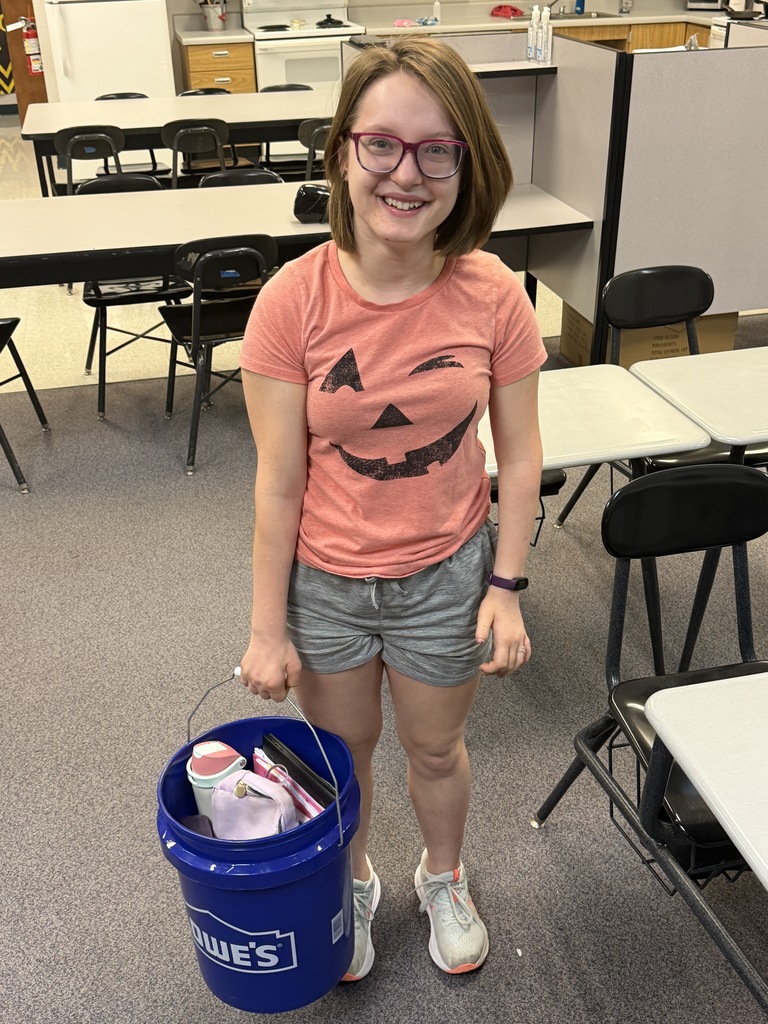 A young person wearing a pink shirt with a pumpkin design holds a blue bucket with a lid.