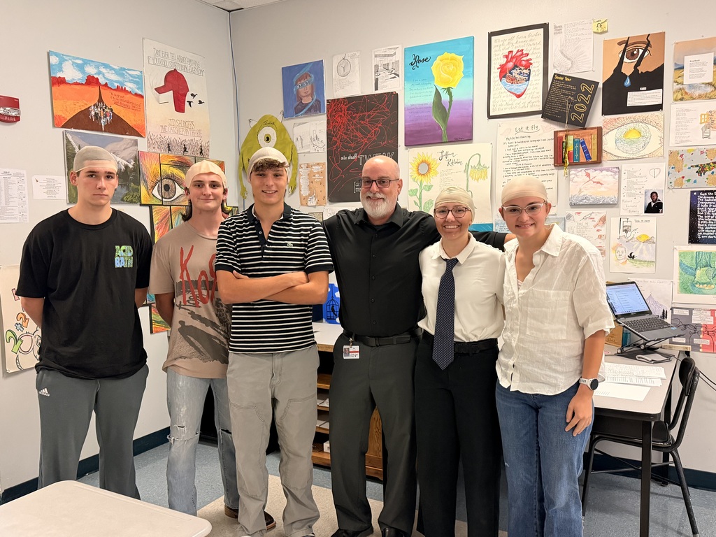 Five people stand in an art room with white walls and multiple paintings and posters.