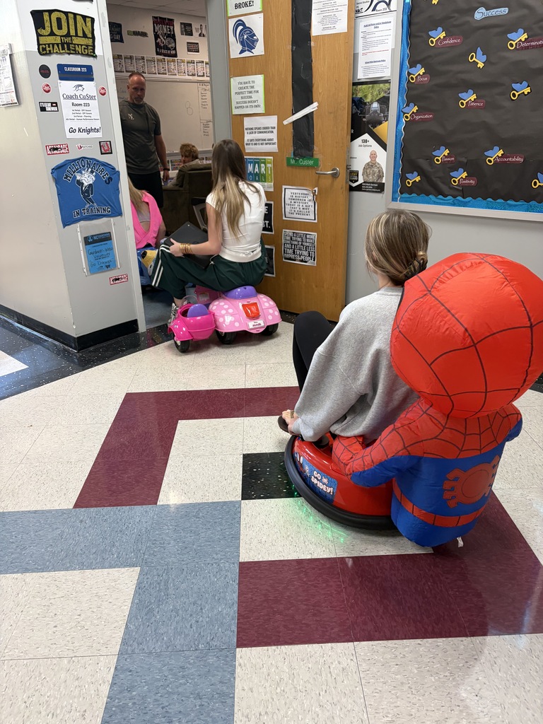 Two children sit on a Spider-Man toy in a hallway with a patterned floor. One child wears a gray sweater.