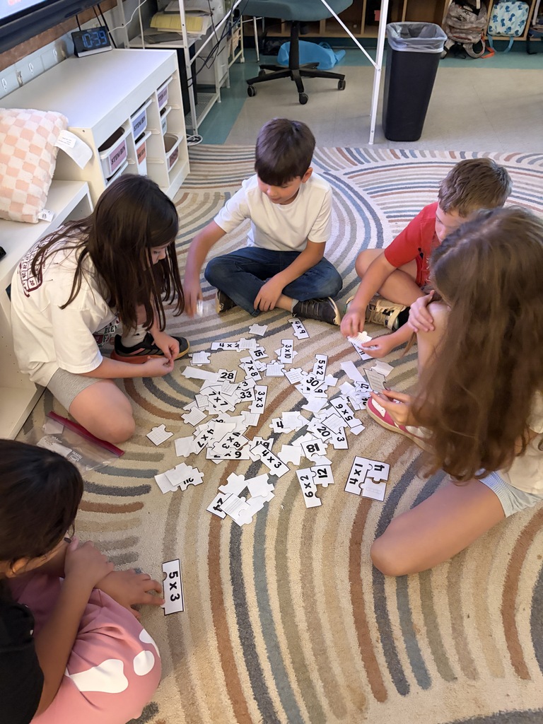 Children are sitting on a carpet and sorting dominoes. They are in a classroom. A white shelf and a trash bin are in the background.
