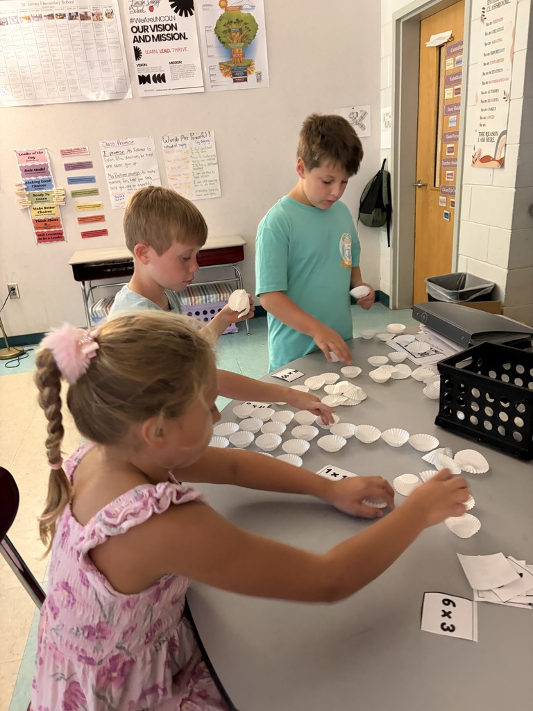 Three children are sitting around a table with white objects and papers on it. They are looking at each other.