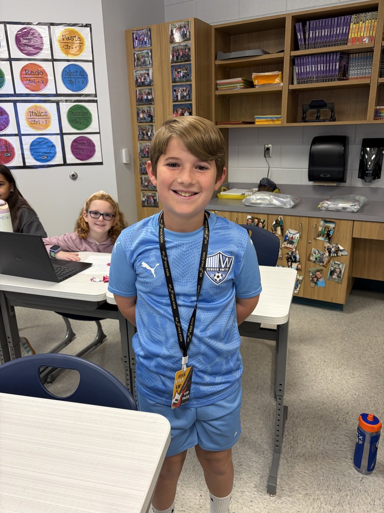 A boy stands in a classroom wearing a blue shirt and shorts, smiling. Behind him, two desks and two children seated. A wooden cabinet, a laptop, and a bottle are in the background.