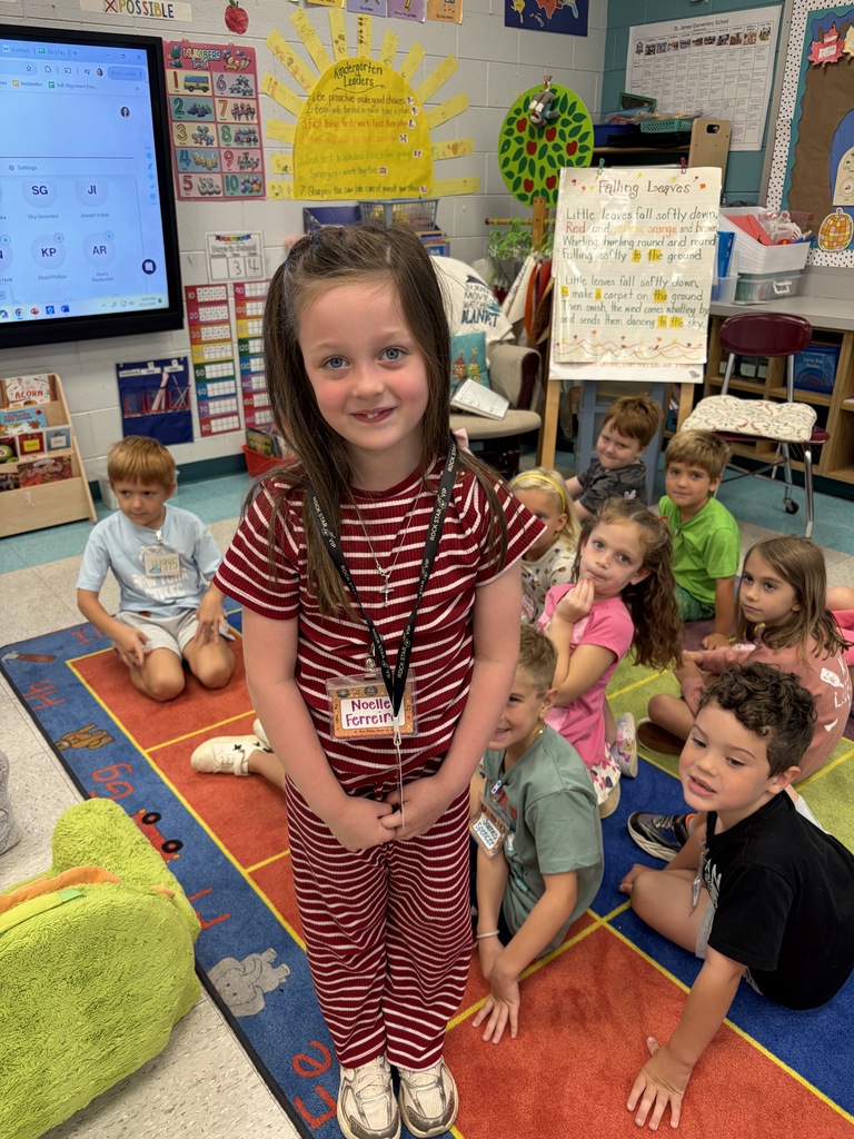 Child stands in front of classroom with young students. Child wears red and white striped shirt. Brightly colored rug on floor.