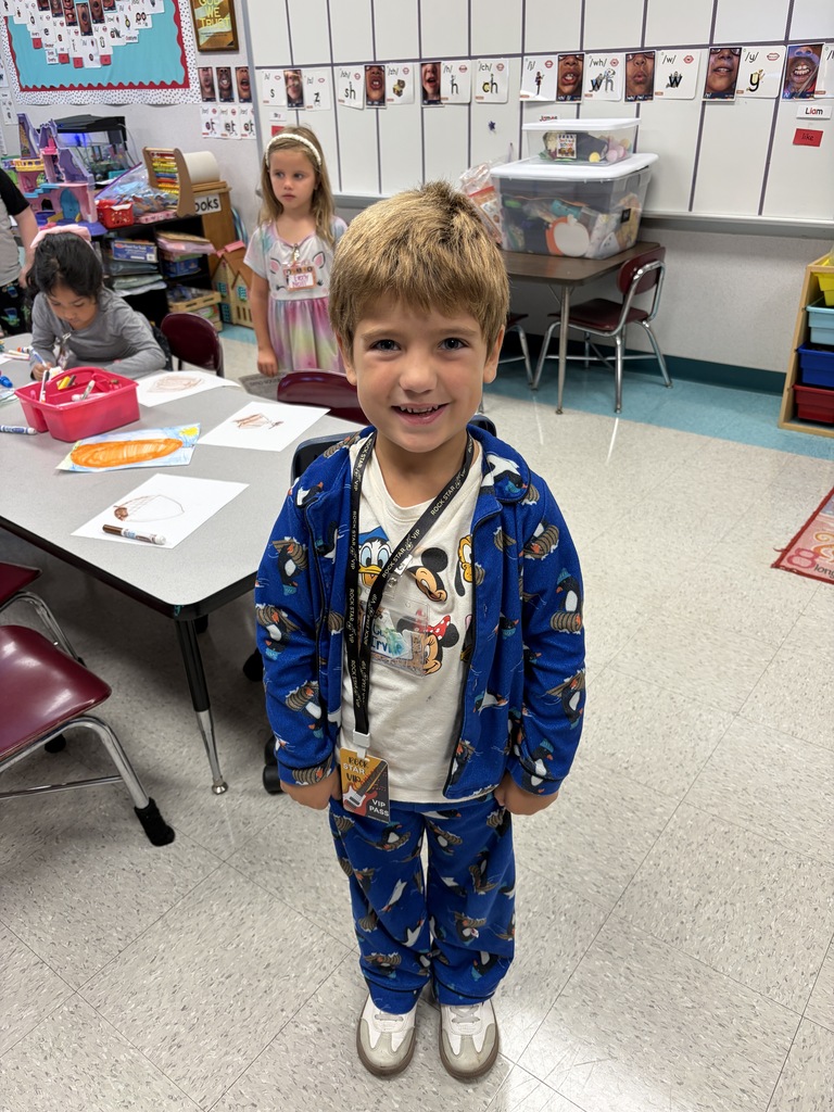 A boy in blue pajamas with cartoon characters stands in a classroom. Behind him is a table with papers.