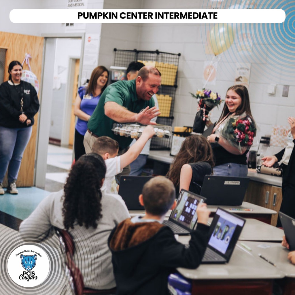A classroom filled with children and adults. A teacher holds a cake. Students use laptops and some hold tablets.