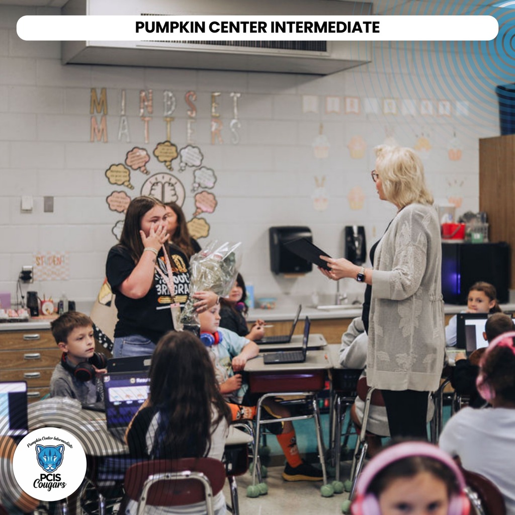 A classroom with children seated at desks, one holding a pumpkin. A woman stands in front of them.