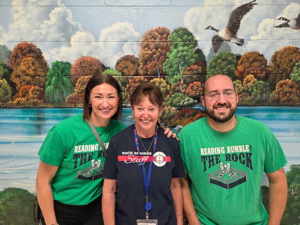Three people smile in front of a backdrop showing trees, a lake, and birds in the sky.