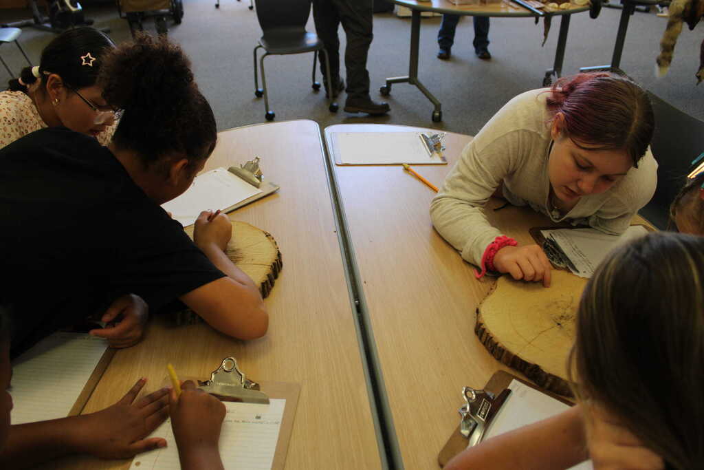 Four individuals seated around a table, engaged in work, with one leaning forward. They have papers and tools on the table.