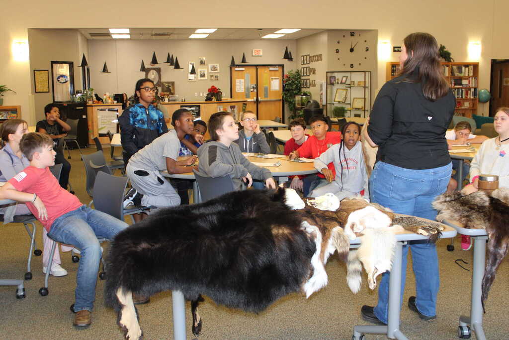 Several individuals are seated around tables, examining animal pelts. A woman stands before them, explaining. Background includes a room with tables and chairs.