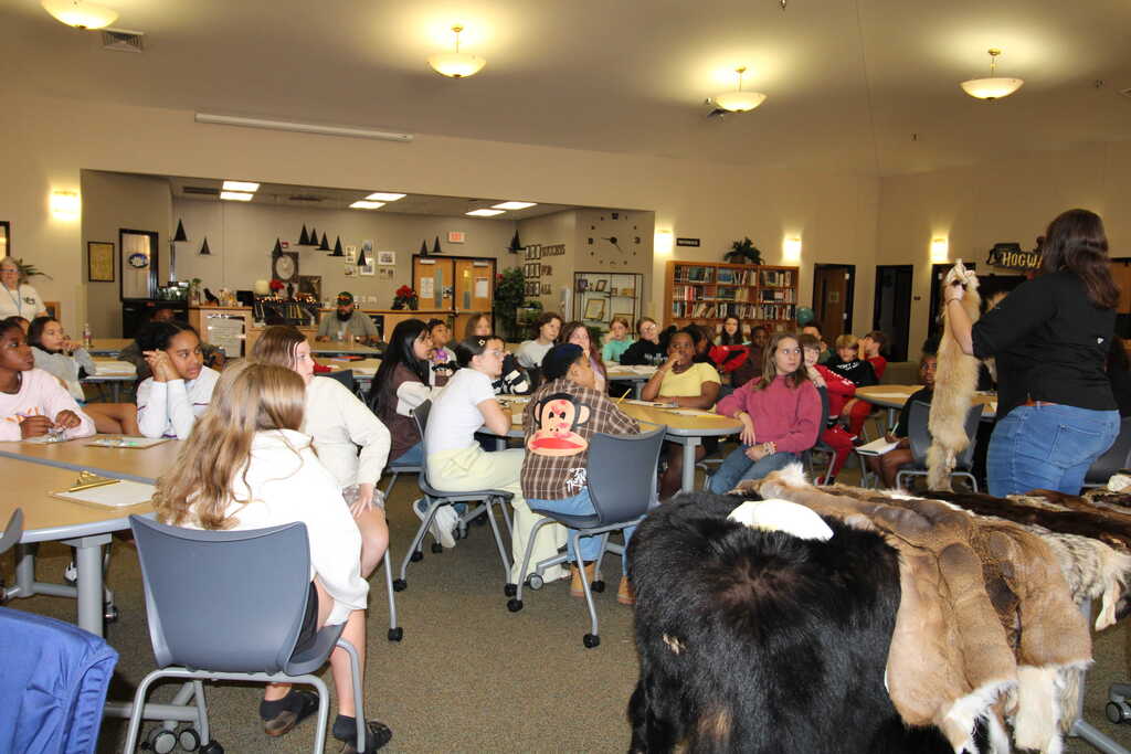 Room with tables, people seated, animal fur on the floor, and a person holding a stuffed animal.