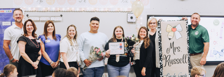 People stand in a classroom, smiling and posing for a photo. A woman holds a certificate and bouquet.