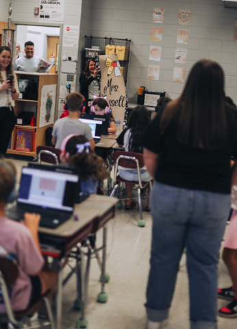 Classroom with students seated at desks using laptops. An adult stands before a group of students.