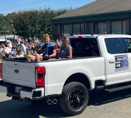 Two people in a pickup truck, one wearing a football jersey, riding in a parade.