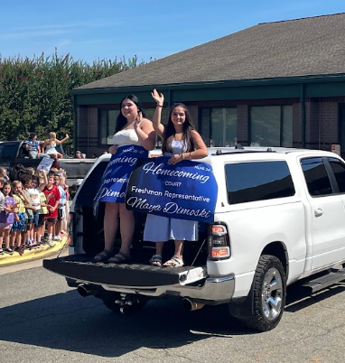 Two women in white dresses wave from a white truck bed. Blue banners are displayed. A building and crowd are behind them.