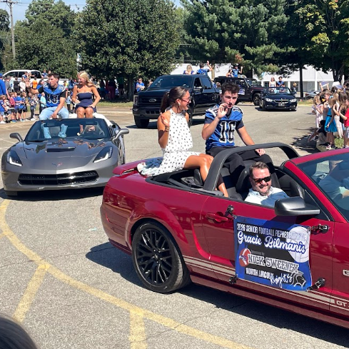 Cars in a parade with people in convertibles, one with a banner, surrounded by a crowd.