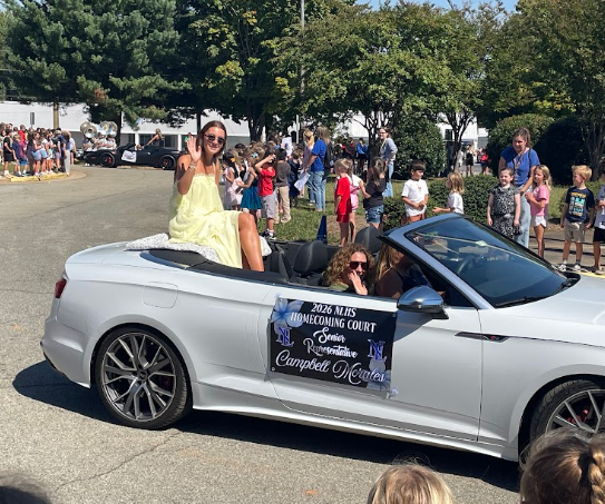 Convertible car with people seated, one wearing yellow, at parade with onlookers.