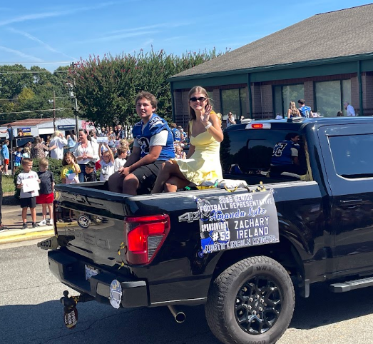 Two people sit in the back of a pickup truck. A large crowd of people stand in the background.