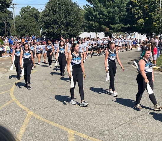 A group of colorguard members in matching uniforms, walking in a parade. The ground is paved with yellow markings.