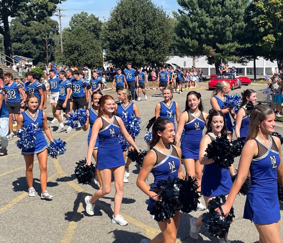Group of cheerleaders in blue uniforms and black pompoms walking in a school parking lot.