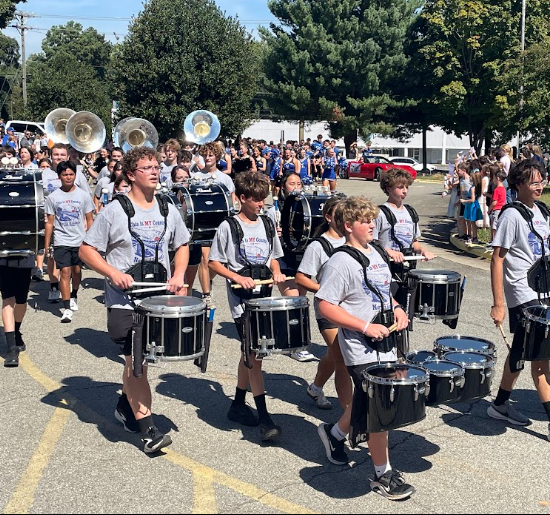 Group of young drummers marching in a line, carrying drums. People and trees in background.