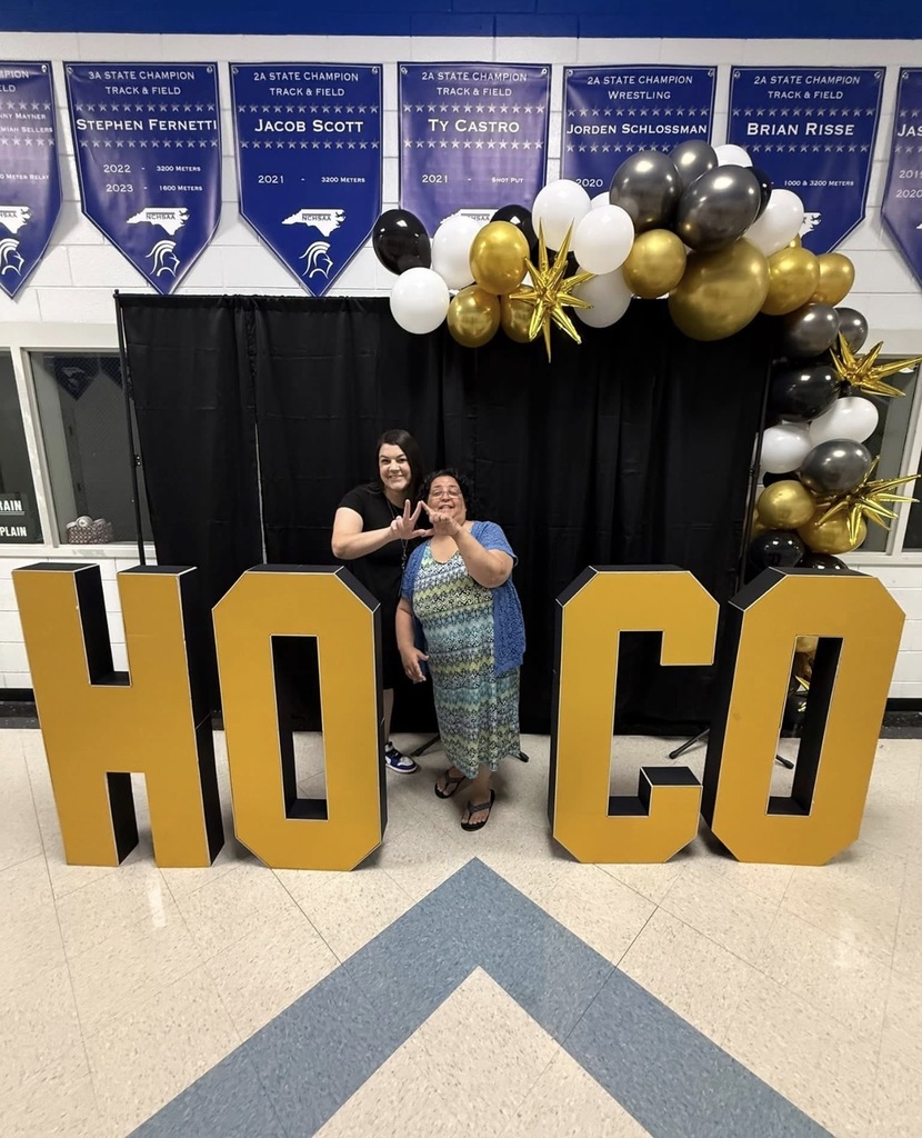 Two women stand behind a large "HOCO" sign, with a black curtain and balloon arch in the background.