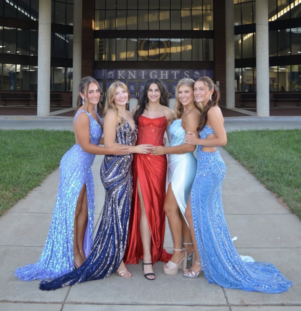 Five women stand close together on a sidewalk in front of a building with pillars. They wear formal gowns.