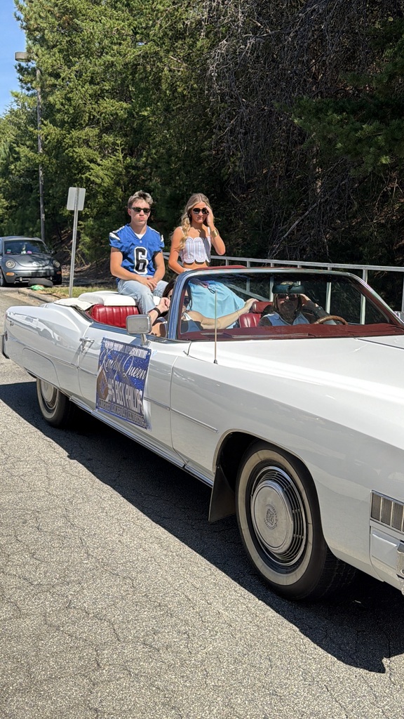 Two people in a white convertible, one wearing a blue jersey, ride through a street lined with trees.