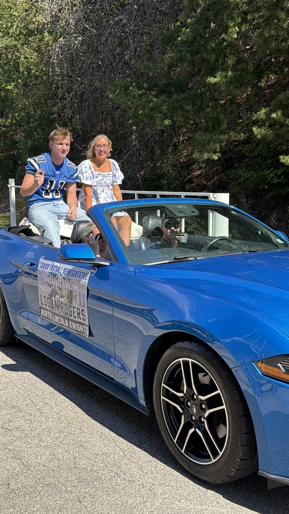 Two people sit in a blue convertible car, one wearing a football jersey, while a banner reads "Lodgers." Trees in the background.