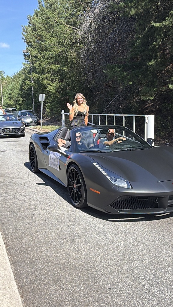 A black sports car drives on a road with two men in the driver and passenger seats. A woman stands on the back, raising her hands. Trees line the road.