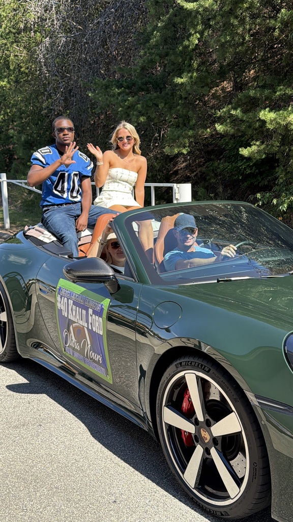 Three people in a green sports car. One person in the back wears a football jersey and sunglasses. Another in the front wears a white dress and waves. The driver wears a blue hat.