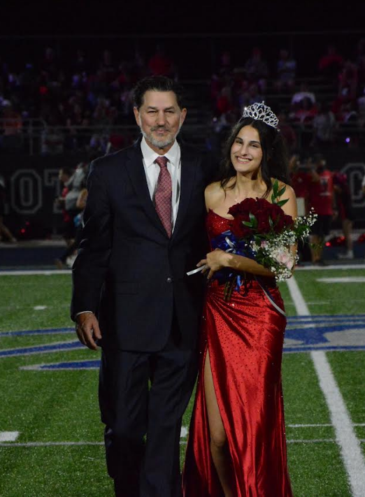 A man in a suit walks with a young woman in a red gown holding flowers on a football field.