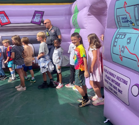 Children and an adult stand in a line around an inflatable smoke alarm structure. Some are looking at the design.