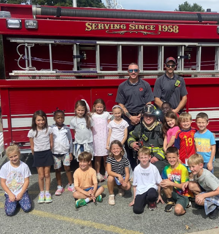 Firefighters in uniform pose with a group of children, all in front of a red fire truck.