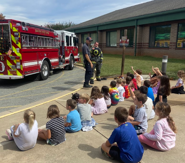 A group of children sit on the sidewalk, watching a firefighter near a fire truck. Behind, a building with glass windows and green trim.