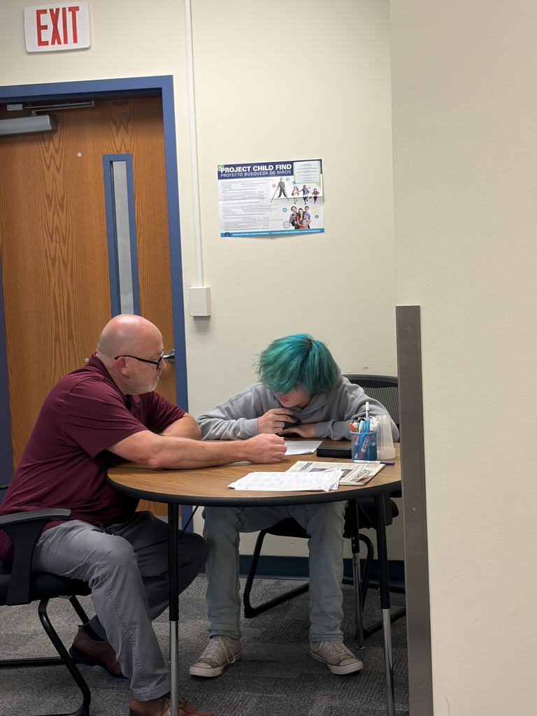 Two individuals seated at a small table. A man and a woman. Papers, pens, and books are on the table.
