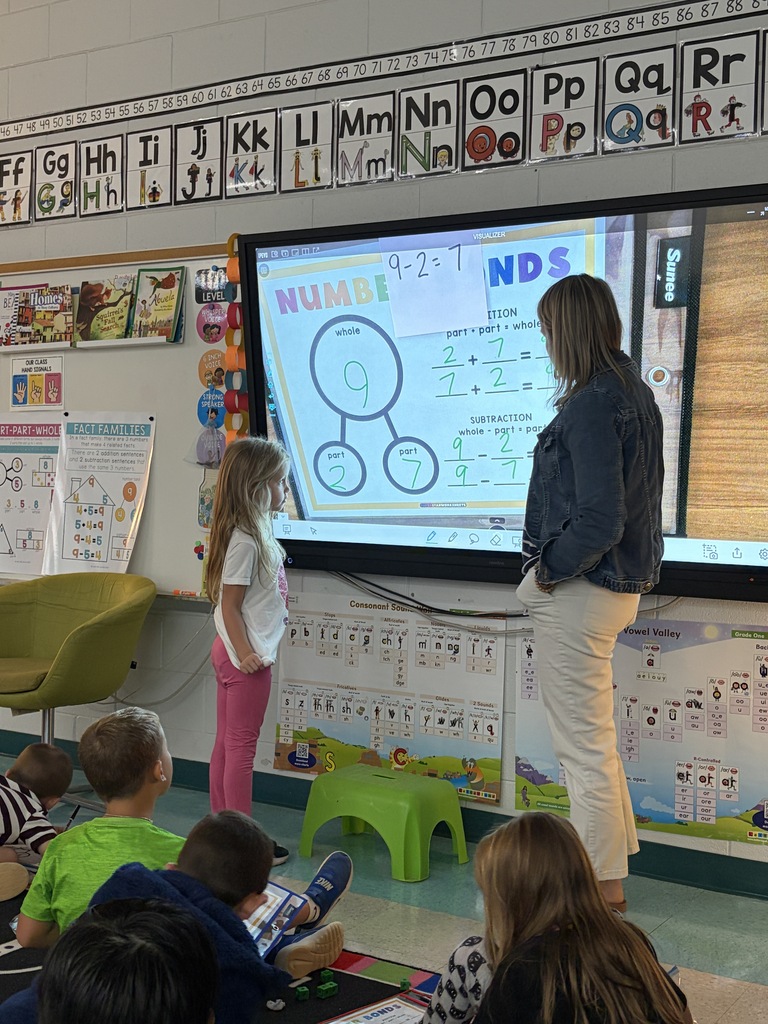 A teacher and a student stand before a classroom screen. Several children sit on the floor, watching.
