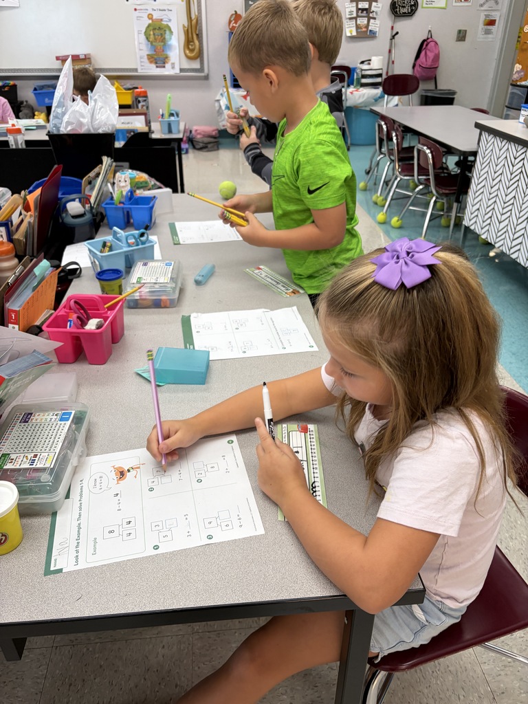 Two children at a desk inside a classroom. A girl in white sits with a pen, focused on paper. A boy in green stands behind her, holding a yellow object.