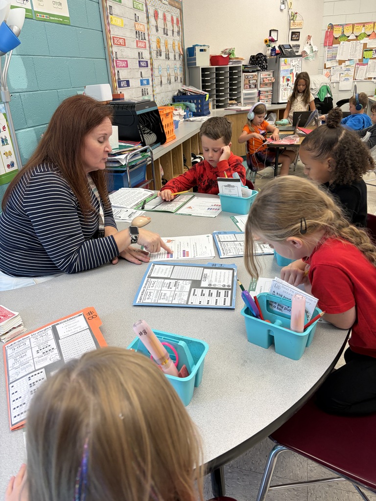 Classroom with several children seated at a table, a woman in front of them, and a teacher interacting with them.