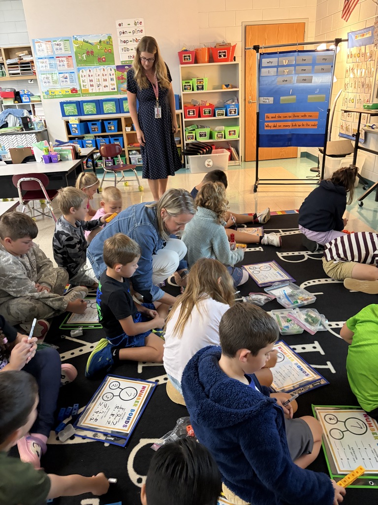 In a classroom, children sit on the floor, studying a book while a woman helps them.