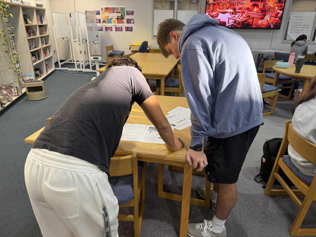 Two people focus on paper on a table in a room with bookshelves, chairs, and a TV.