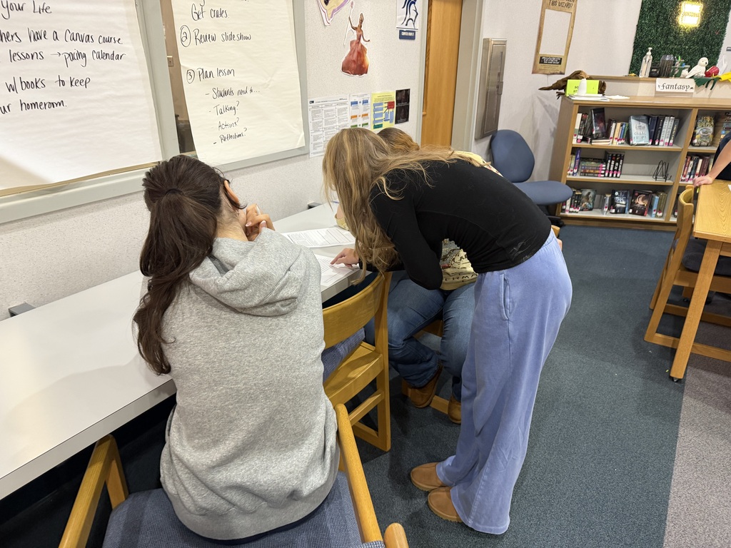 Two people in a classroom, one sitting at a desk, the other standing and looking over their shoulder.