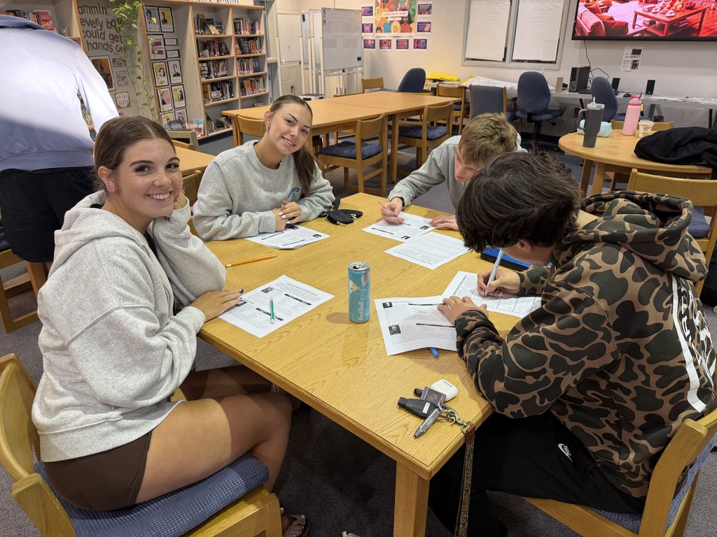 A group of students sit at a table in a classroom, with papers and pens on the table.